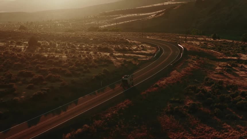 An aerial view captures a camper van driving along a winding road through a scenic, sun-drenched rural landscape at golden hour