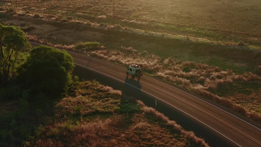 An aerial view follows a camper van on a scenic road trip driving alongside a river through the golden hour countryside