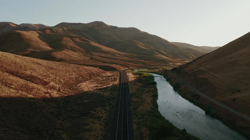 A tranquil aerial view follows an empty road and a peaceful river through a vast mountain canyon during golden hour