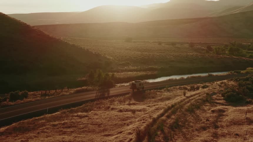 An aerial view follows a camper van driving on a scenic country road through a golden, sun-drenched valley at sunset