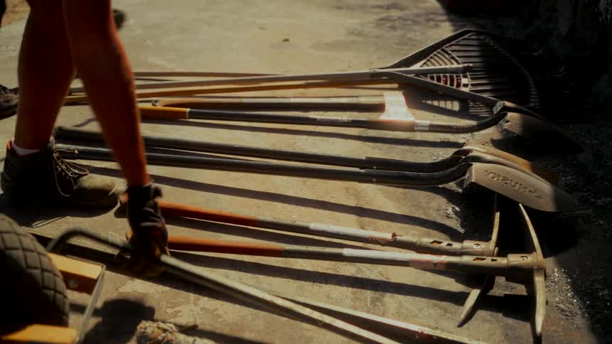 A worker in boots and gloves lays down a crowbar next to a collection of shovels, rakes, and pickaxes on a concrete floor