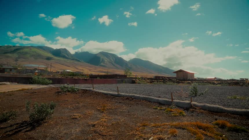 A wide, static shot of a rural landscape featuring majestic green mountains under a bright blue sky with fluffy white clouds