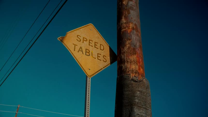A weathered yellow 'Speed Tables' road sign is attached to a rustic wooden utility pole against a clear, deep blue sky