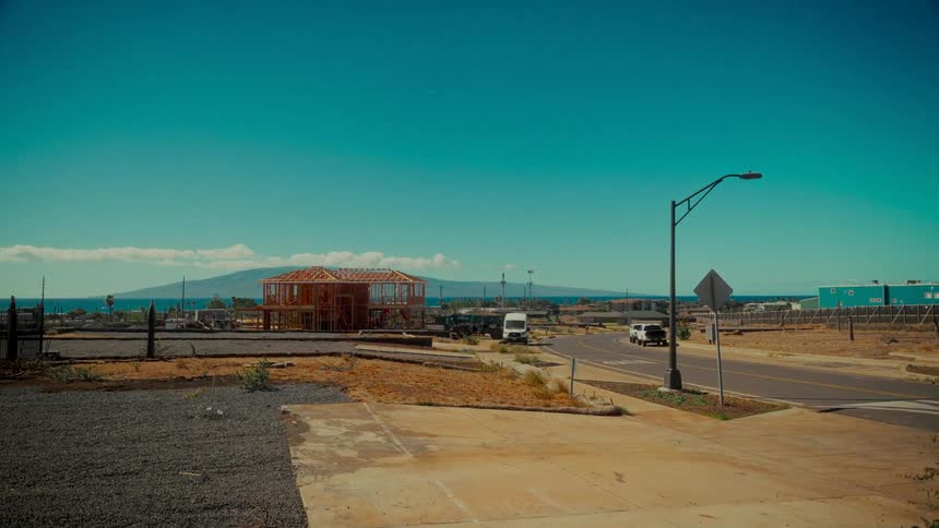 A new home is being framed in a desolate, sun-drenched coastal landscape with an island visible across the ocean