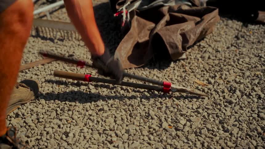 A gloved hand picks up a pair of large pruning shears from the gravel in slow motion