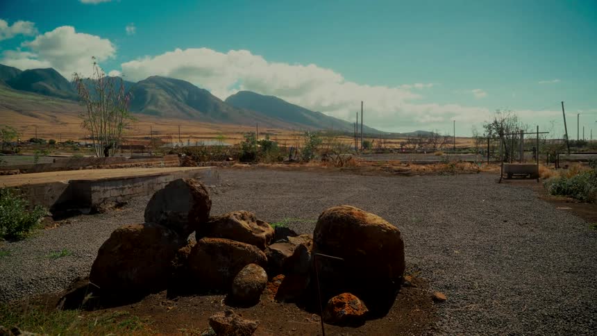 An empty gravel lot with large rocks sits in a dry, desolate valley with mountains in the background