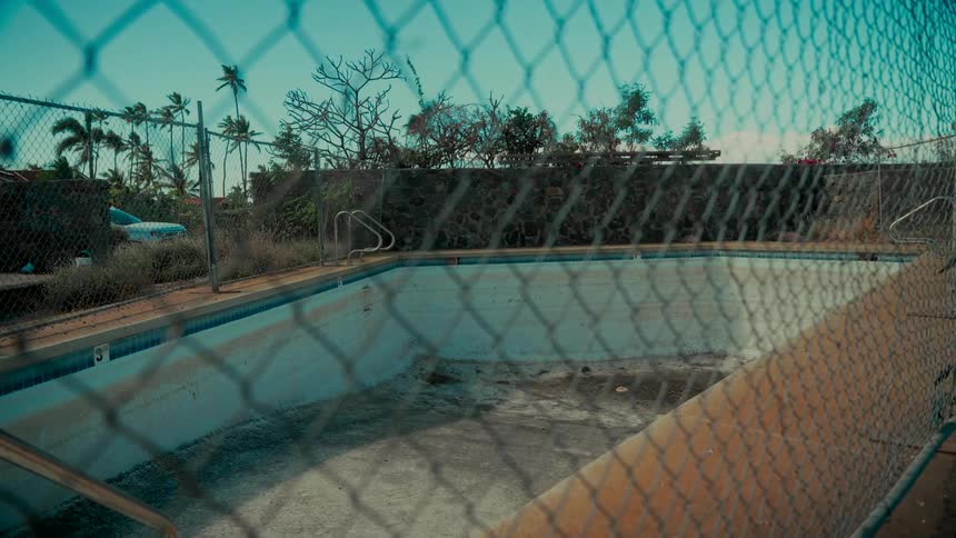 An eerie static shot of an empty and abandoned swimming pool seen through a chain-link fence on a sunny day