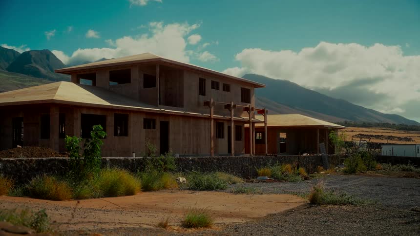 The wooden frame of a large, custom home under construction stands against a backdrop of mountains on a partly cloudy day