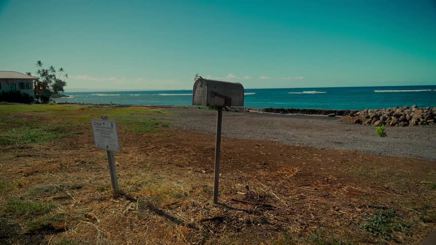 An old, weathered mailbox stands alone in a coastal field overlooking the bright blue ocean on a sunny day