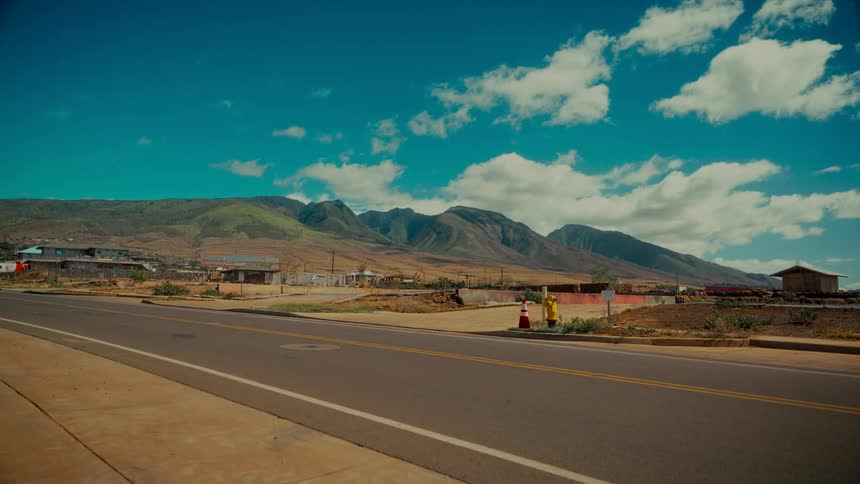 An empty road runs through a desolate town at the foot of barren mountains under a partly cloudy sky