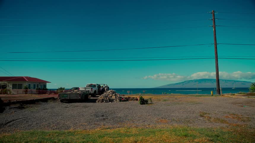 A wide static shot of a coastal gravel lot with a pickup truck and trailer, overlooking the ocean and a distant island under a clear blue sky