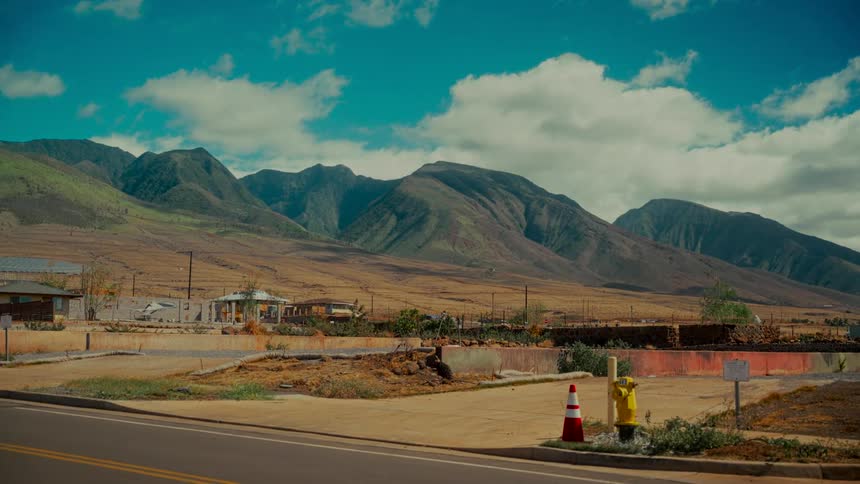 The desolate aftermath of a wildfire in Lahaina, with destroyed homes and barren land set against the backdrop of the West Maui Mountains as a car drives by