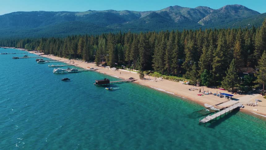 An aerial view pulls back over the turquoise waters of a mountain lake, revealing a sandy shoreline dotted with piers and backed by a dense pine forest