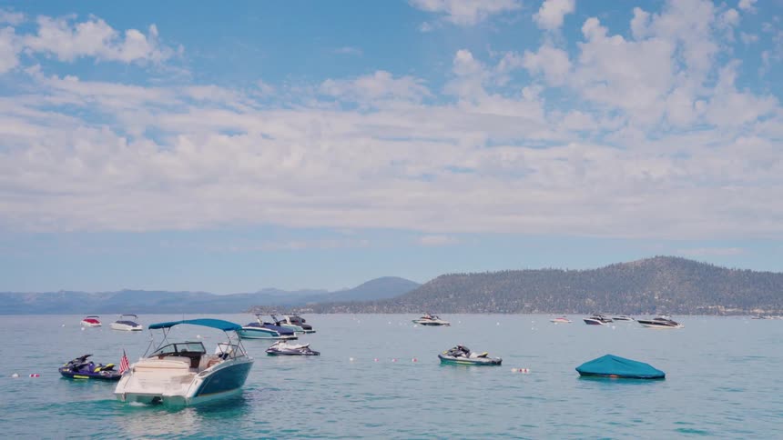 A wide shot of numerous boats and jet skis floating on a calm lake on a sunny day with mountains in the background