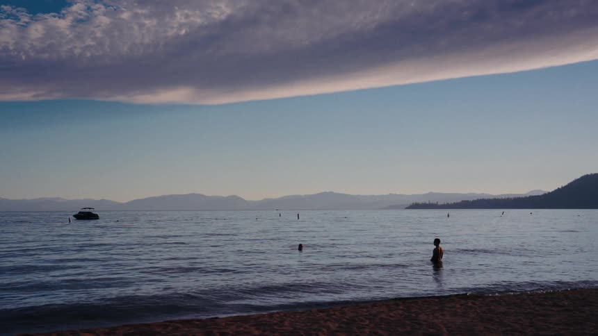A serene view of people wading in a calm lake at dusk, with mountains in the distance and a dramatic cloud overhead