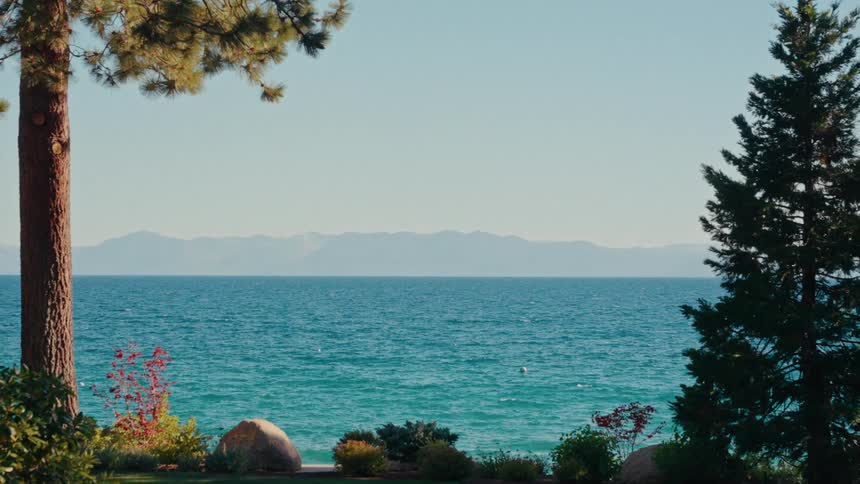 A scenic, static shot of a vast blue lake with distant mountains, framed by shoreline trees on a clear day
