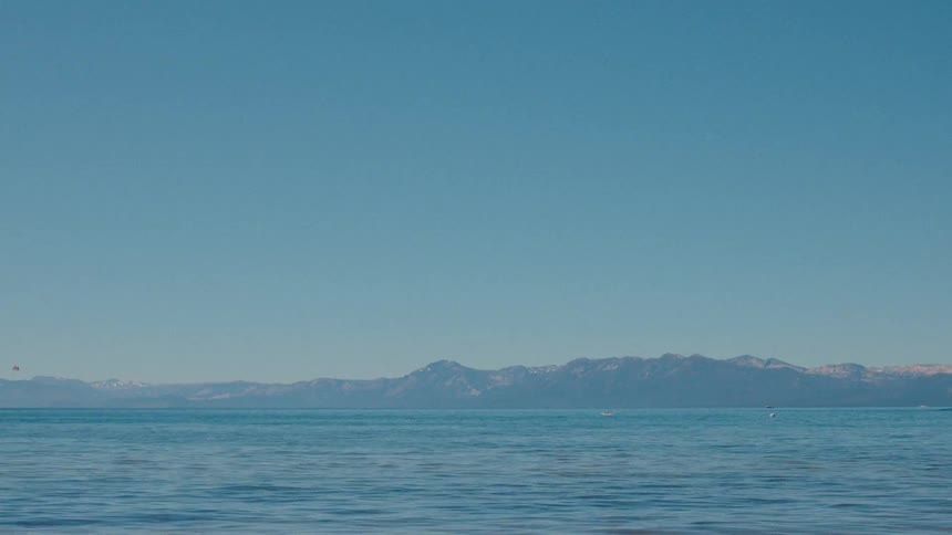 A peaceful, wide view of a calm blue lake against a backdrop of distant, snow-capped mountains under a vast, clear sky