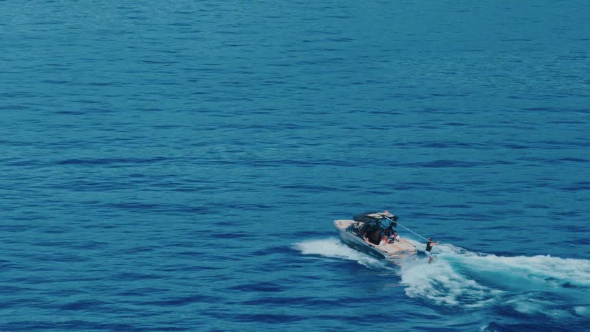 An aerial slow-motion view shows a person wakeboarding behind a speed boat on the vast blue sea