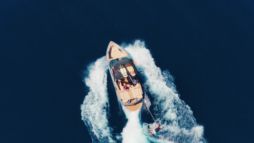 An aerial top-down shot shows a man wakeboarding behind a speedboat, carving through the blue ocean before eventually falling into the water