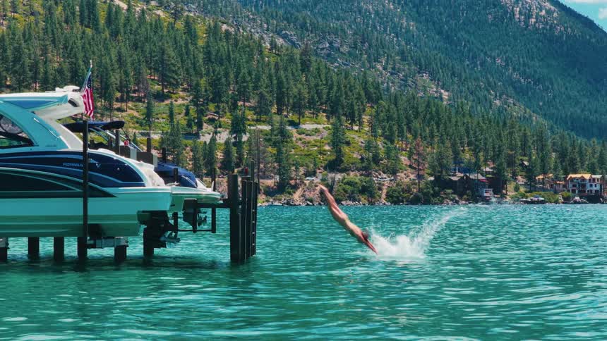 A young couple jumps and dives into a clear turquoise mountain lake from a dock in slow motion