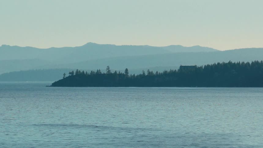 A tranquil and hazy morning view of the calm waters of a vast lake with distant layers of mountains