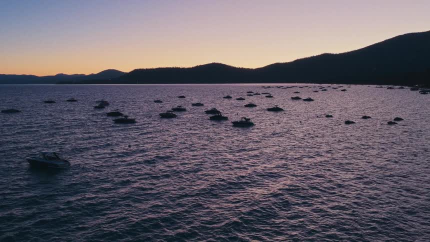 Boats are anchored on a calm lake as the sun sets behind silhouetted mountains