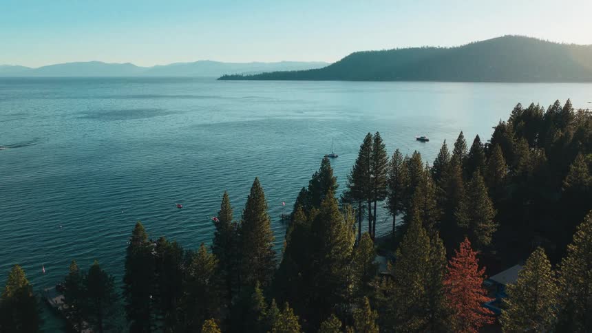 An aerial view of the serene Lake Tahoe at sunrise, with a lush evergreen forest along the shoreline and boats on the water
