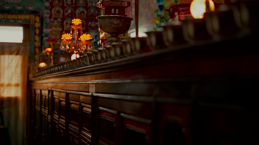 A tranquil dolly shot reveals a long row of traditional offering bowls on a wooden altar inside a warmly lit Buddhist monastery