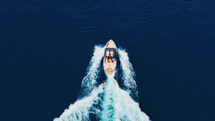 Top-down aerial footage of a wakesurfer being towed by a motorboat across the vast, deep blue ocean