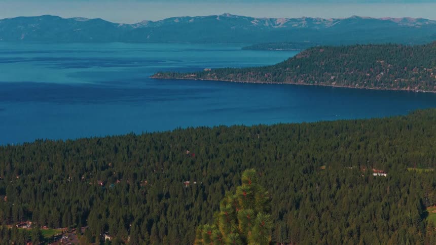 A vast and beautiful slow zoom-out shot reveals the stunning blue waters of Lake Tahoe, surrounded by a dense evergreen forest and distant mountains