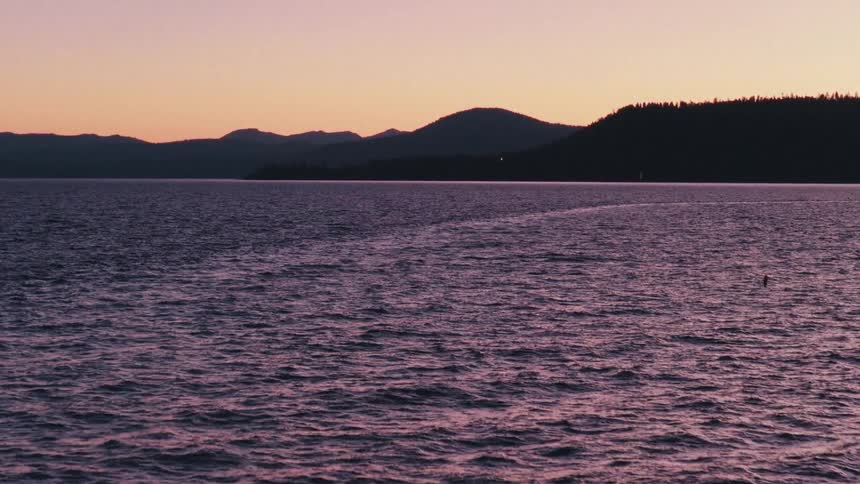 The calm, purple waters of a vast lake ripple gently at dusk, with the silhouette of distant mountains against a soft sunset sky