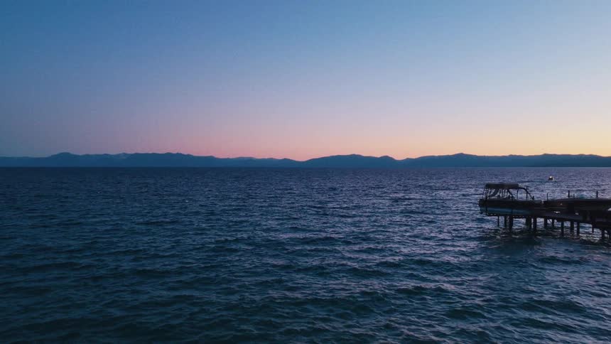 A tranquil evening scene overlooking a vast lake, with a wooden pier and distant mountains silhouetted against the sunset sky