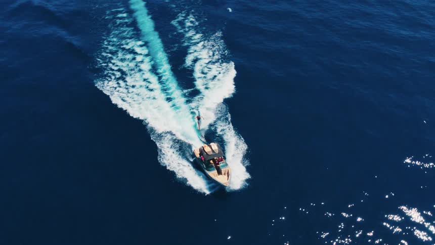 Top-down aerial footage tracks a motorboat pulling a wakesurfer across the shimmering blue ocean on a sunny day