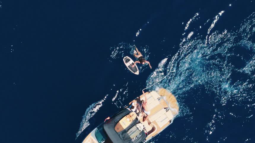 An aerial top-down slow-motion shot of a person successfully getting up on a wakeboard behind a motorboat in the deep blue ocean