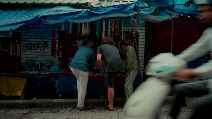 Three friends browse for handmade jewelry and trinkets at an outdoor street market stall as a scooter passes by