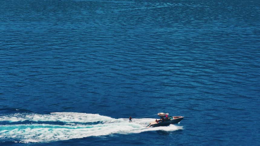 A high-angle slow-motion shot follows a water skier gliding across a stunningly blue lake, pulled by a speedboat under a clear sunny sky