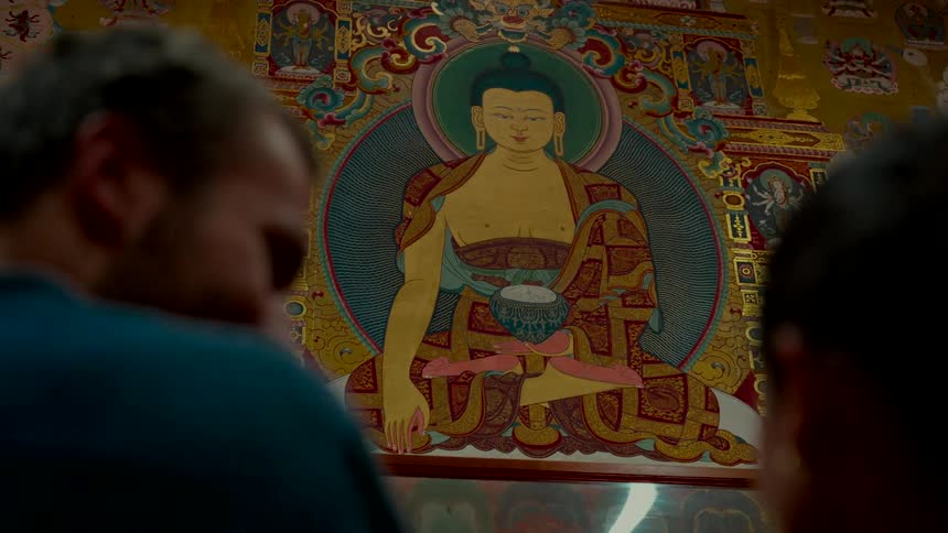 An over-the-shoulder shot captures two people as they admire a large, colorful, and traditional painting of the Buddha inside a temple