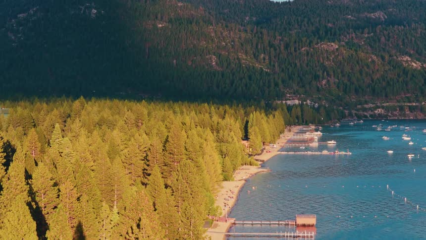 Golden hour sunlight illuminates the pine forest and beach along the shore of a beautiful mountain lake where people are relaxing