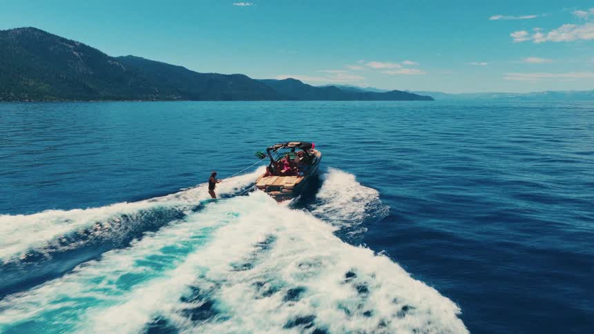 A wakesurfer rides the waves behind a motorboat full of friends on a scenic blue lake surrounded by mountains