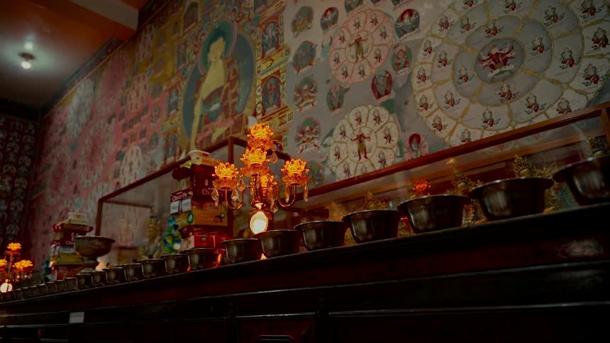 A row of offering bowls and a glowing golden lamp sit before a large, colorful mural inside a Buddhist temple