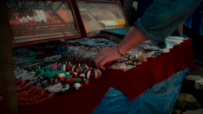A traveler's hand carefully selects a traditional beaded necklace from a vibrant market stall display