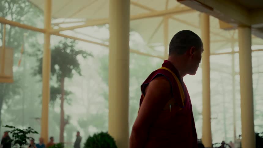 A Tibetan monk in traditional robes stands inside a temple hall as fog rolls in outside