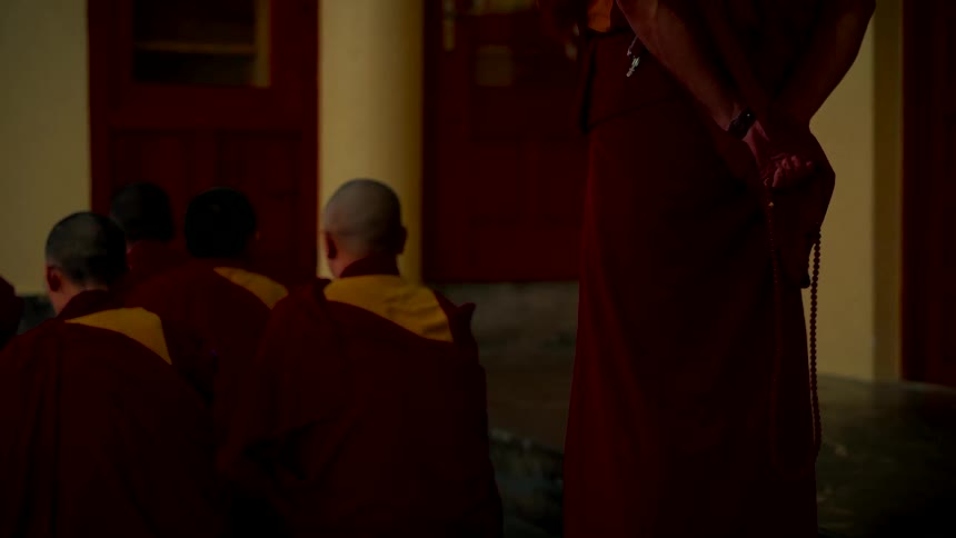 A standing monk holds prayer beads behind his back while watching over a group of meditating monks in a quiet, dimly lit temple