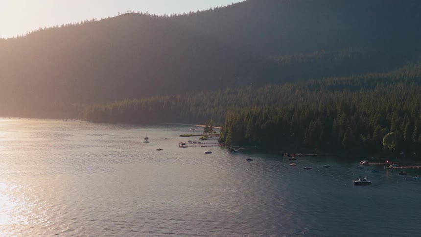 An expansive aerial shot reveals a beautiful mountain lake cove with numerous boats floating on the calm water during a hazy, golden sunset