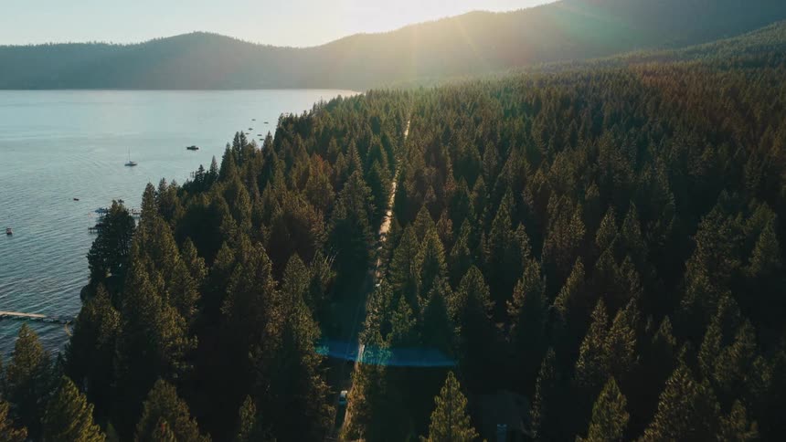 A stunning aerial view captures a car driving through a dense pine forest along a lakeshore as the sun sets over the mountains