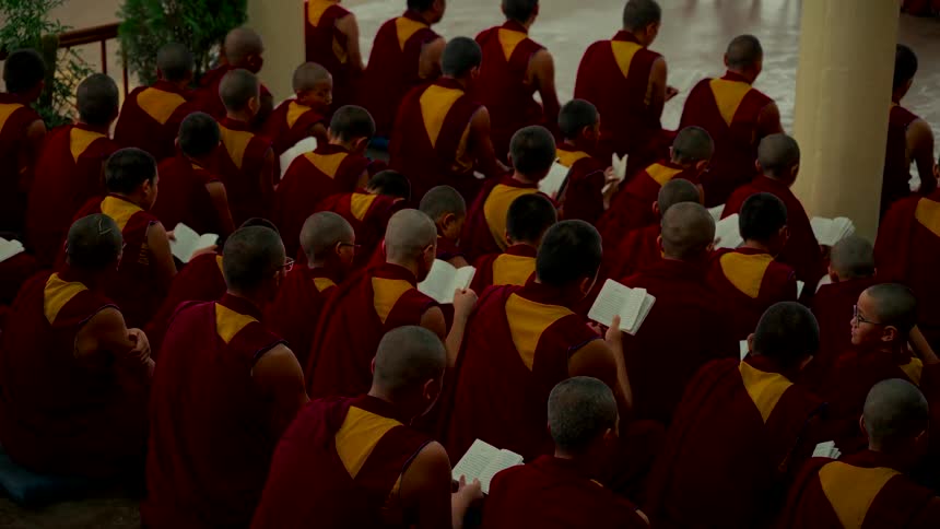 From above, a large group of Buddhist monks in red and yellow robes sit on the floor of a monastery reading from their prayer books