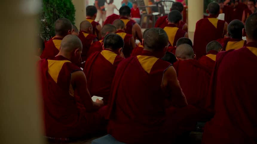A large group of Buddhist monks with shaved heads and red robes sit together during a religious ceremony at a monastery