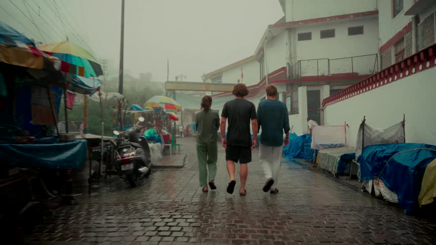 Following from behind, three friends walk down a wet, foggy street lined with market stalls in an Asian village