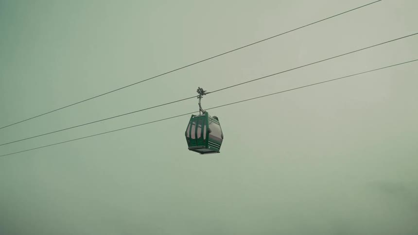 A lone green cable car glides through the thick fog on a cable line as the camera slowly zooms out