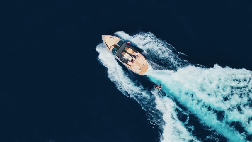 Aerial top-down slow-motion view of a person wakeboarding behind a luxury motorboat on the open ocean